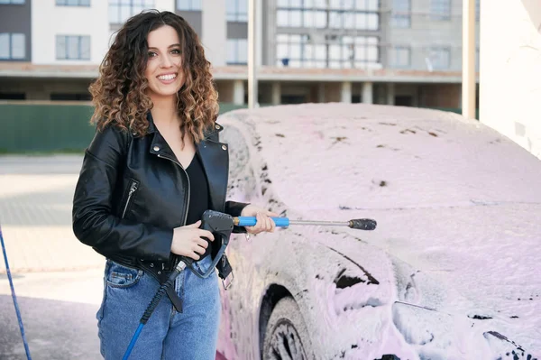 Cheerful young woman in leather jacket looking at camera and smiling while holding car washing tool. Beautiful stylish woman with car cleaning gun standing by automobile covered with foam.