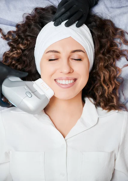 Top view of beautiful young woman with closed eyes wearing white headband while receiving facial treatment at wellness center. Cheerful female patient having face lifting procedure and smiling.