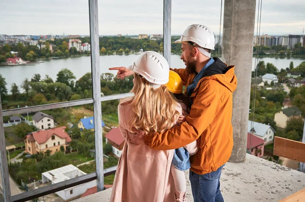 父母带着女儿站在在建公寓楼内的背景图 男人和女人带着孩子戴着建筑用的头盔 从未来的家欣赏城市美景 — 图库照片