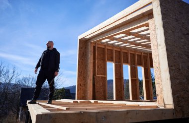 Male builder building wooden frame house. Bald man standing on construction site, inspecting quality of work on sunny day with blue sky on background.