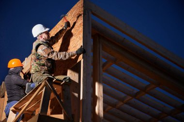 Carpenters hammering nail into OSB panel on the wall of future cottage in the evening. Men workers building wooden frame house. Carpentry and construction concept.
