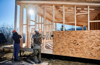 Men workers building wooden frame house on pile foundation. Back view of carpenters using tape measure for measuring wooden planks. Carpentry concept.