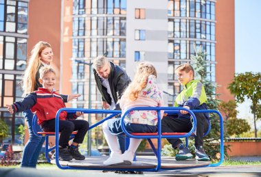 Mutlu aile - baba, anne ve çocuklar şehrin yüksek binalarının modern avlusunda birlikte eğleniyorlar. Çocuklarla oynayan gülümseyen ebeveynler, çocuk parkında sallanan.