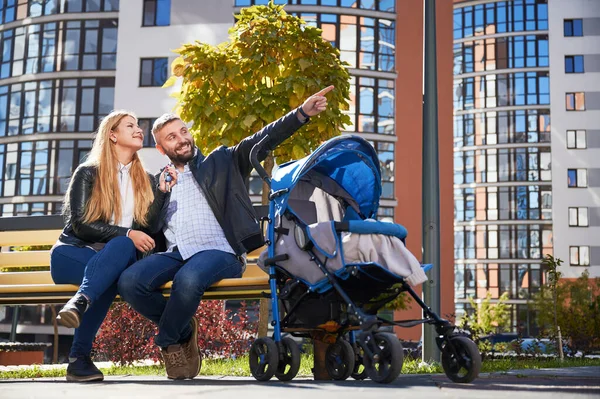 快乐的一家人一起坐在蓝色婴儿车里滑行 微笑的女人拿着钥匙 男人指着新城区的新公寓 背景的现代住宅 — 图库照片