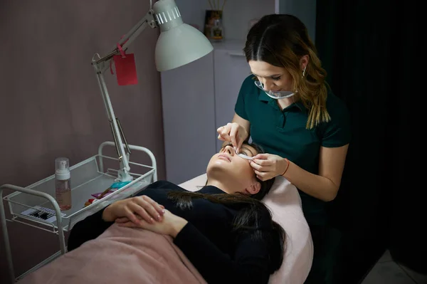 Female beauty specialist in protective face mask placing eye patch on client face while woman lying on daybed. Eyelash stylist preparing woman eyes before applying lashes extensions.
