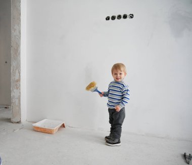 Sevimli küçük çocuk elinde fırça tutarken kameraya bakıyor ve gülümsüyor. Elinde boya aletleri olan neşeli bir erkek çocuk apartman dairesinde beyaz duvara karşı duruyor..