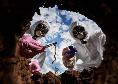 Koruyucu tulumlu ve gaz maskeli araştırmacıların toprak ve sakinlerini incelediği iç kuyuya bakın. Bir laboratuvar asistanı cımbızla toprak solucanı, diğeri de sıvı cam matara tutuyor..