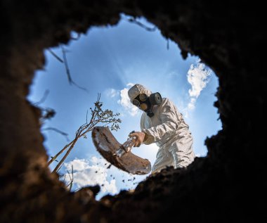 Çevre bilimci, çukur kazıyor, kürek ve ağaç dikiyor, gaz maskesi takıyor ve açık havada koruyucu tulumlar giyiyor, mavi bulutlu gökyüzünün arka planında..
