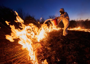 İtfaiyeci arka planda mavi gökyüzü olan yangını söndürüyor. Koruyucu radyasyon kıyafeti giyen ve gaz maskeli bir adam yanan çimlerin üzerine dumanla su döküyor. Doğal afet kavramı.