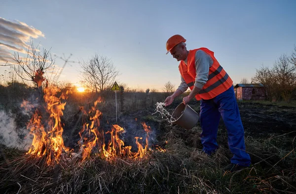 Fireman ecologist extinguishing fire in field with warning sign with exclamation mark end evening sky on background. Male environmentalist holding bucket and pouring water on burning dry grass.