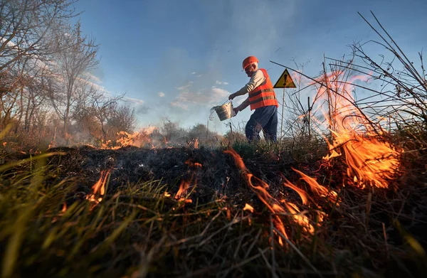 Firefighter ecologist extinguishing fire in field with evening sky on background. Male environmentalist holding bucket and pouring water on burning dry grass near warning sign with exclamation mark.
