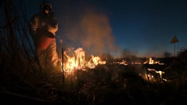 夜空を背景に戦場で消火活動 保護放射線スーツの男とガスマスク煙と燃える芝生の上に水を注ぐ 自然災害の概念 — ストック動画