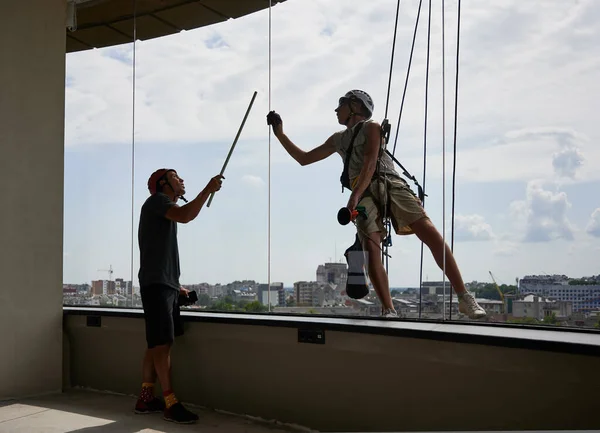 Industrial mountaineering professional cleaner hanging on rope and wiping window with rag while colleague standing inside building, pointing on the dirty window.