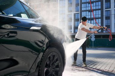 Şapkalı bir adam siyah arabasını temizlerken yüksek basınçlı su kullanıyor. Konut binasının arka planında akan suyla oto yıkama.