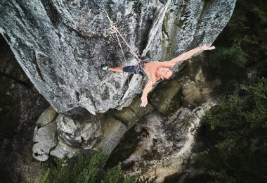 Genç adam iple yüksek kayaya tırmanıyor. Korkusuz dağcı rota zirvesinde başarıyı kutluyor. Ekstrem spor, macera ve aktif yaşam tarzı kavramı. Hava görünümü.