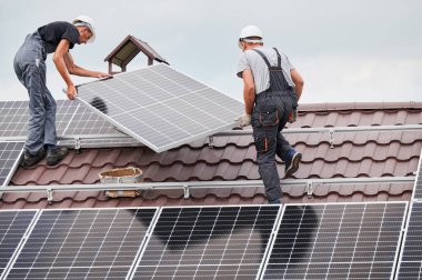 Erkek teknisyenler fotovoltaik güneş modüllerini evin çatısına kaldırıyorlar. Kasklı işçiler dışarıda güneş paneli sistemi kuruyor. Alternatif ve yenilenebilir enerji kavramı.