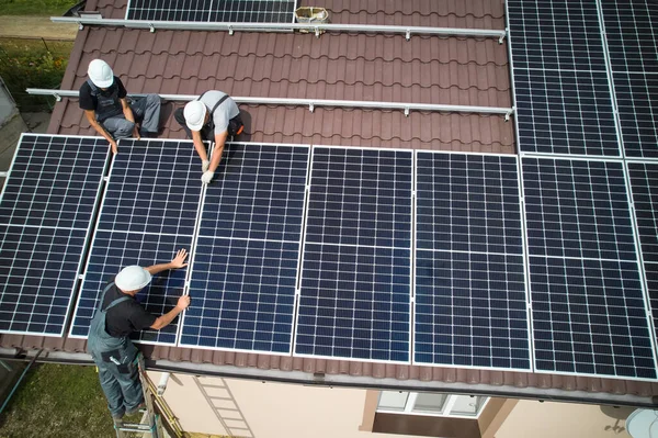 Men technicians mounting photovoltaic solar moduls on roof of house. Electricians in helmets installing solar panel system outdoors. Concept of alternative and renewable energy. Aerial view.