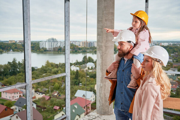 Daughter sitting on fathers shoulders and pointing at something at construction site. Family with child future homeowners observing apartment building under construction.