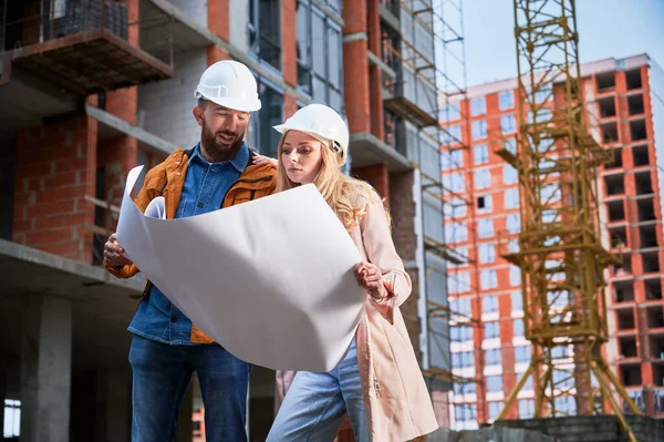 Couple in safety helmets studying architectural drawings outside apartment building under construction. Man and woman discussing building plan outdoors at construction site.