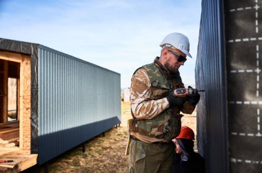 Erkek müteahhit, gelecekteki kulübenin ön cephesi olarak kullanılan siyah kıvrımlı demir çarşaf takıyor. Ahşap çerçeve inşa eden bir işçi. Marangozluk ve inşaat konsepti.