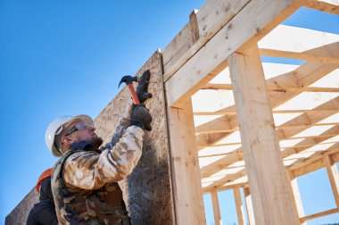 Marangoz, OSB panelinin duvarına çivi çakıyor. Ahşap çerçeve inşa eden bir işçi. Marangozluk ve inşaat konsepti.