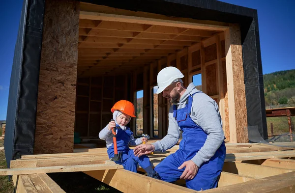 Father with toddler son building wooden frame house. Boy helping his daddy, playing with kid on construction site, wearing helmet and blue overalls on sunny day. Carpentry and family concept.