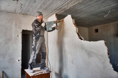 Tadilat sırasında apartman duvarını yıkarken çekiç matkabı kullanan iş elbiseli bir adam. Erkek işçi ahşap masada duruyor ve özel bir aletle duvarı deliyor..