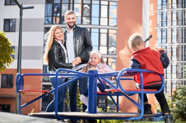 Mutlu aile - baba, anne ve çocuklar birlikte eğleniyorlar. Gülümseyen ebeveynler birbirlerine sarılırken, çocuklar yeni şehir bölgesindeki çocuk parkında sallanıyorlar. Arka planda modern konut binaları.