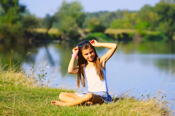 Маленькая девочка, сидящая на берегу реки в летнее время — стоковое фото