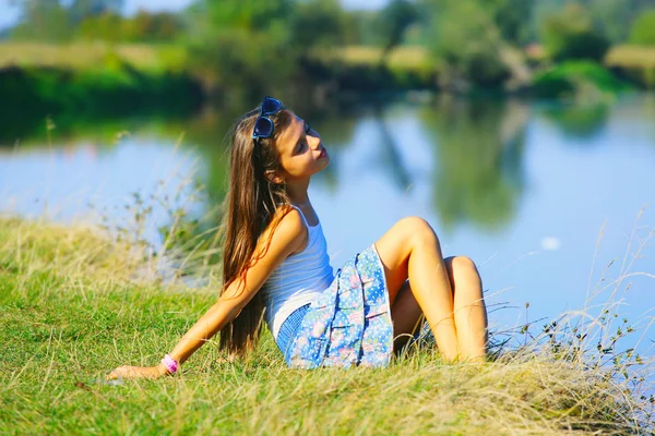 Маленькая девочка, сидящая на берегу реки в летнее время Стоковая Картинка