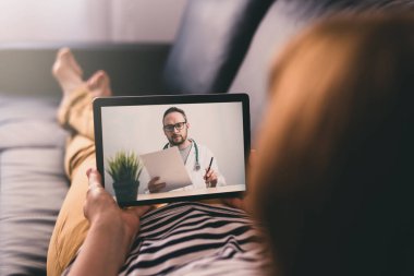 Evdeki kanepede uzanmış dijital tablet kullanarak bir doktorla sohbet eden bir kadın. Teletıp kavramı.