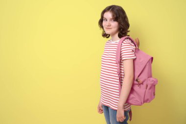 Portrait of a schoolgirl standing, smiling and holding a backpack, looking at camera, on yellow background