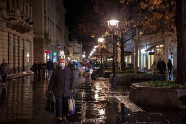 BELGRAD, SERBIA - NOVEMBER 3, 2021: Coronavirus covid 19 krizi sırasında gece elinde alışveriş çantası ve alışveriş arabasıyla solunum maskesi takan yaşlı bir kadının seçici bulanıklığı