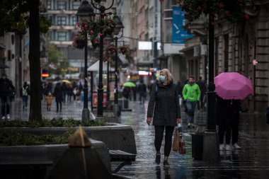 BELGRADE, SERBIA - OCTOBER 10, 2021: Selective blur on an Old senior woman wearing a respiratory facemask walking under rain with a groceries bag and shopping cart during coronavirus covid 19 crisis