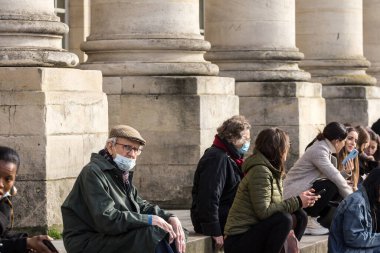 BORDEAUX, FRANCE - FEBRUARY 25 2022: Old senior man wearing a respiratory face mask sittting relaxed in the middle of a crowd in the city center of Bordeaux, during the coronavirus covid 19 health crisis