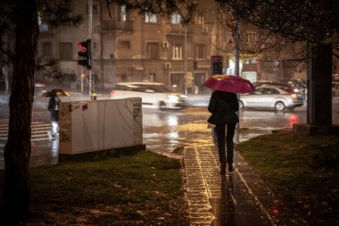 BELGRAD, SERBIA - NOVEMBER 4, 2021: Yağmurlu bir sonbaharda yağmurlu bir akşamda yağmurlu bir sokakta şemsiyeyle yağmurda yürüyen yaşlı bir kadın için seçici bulanıklık arkasında hız bulanıklığı