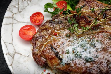 Beyaz bir tabakta tereyağlı, otlu ve kiraz domatesli ızgara biftek, koyu bir arka planda.