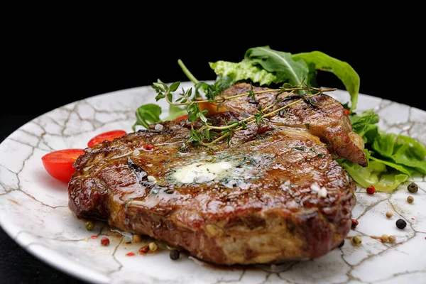 Beyaz bir tabakta tereyağlı, otlu ve kiraz domatesli ızgara biftek, koyu bir arka planda. — Stok fotoğraf