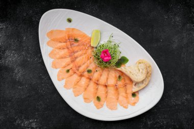 Bitkilerle kaplı somon carpaccio, bir tabakta, koyu bir arka planda.