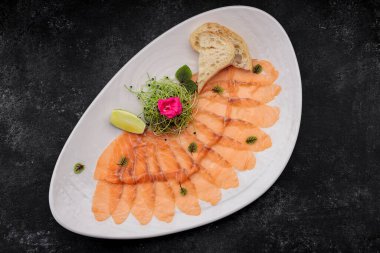 Bitkilerle kaplı somon carpaccio, bir tabakta, koyu bir arka planda.