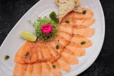 Bitkilerle kaplı somon carpaccio, bir tabakta, koyu bir arka planda.
