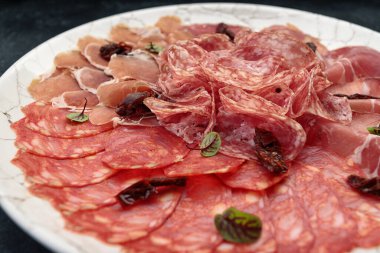 Beyaz bir tabakta dilimlenmiş et, siyah bir arka planda. Sosis, jamon.