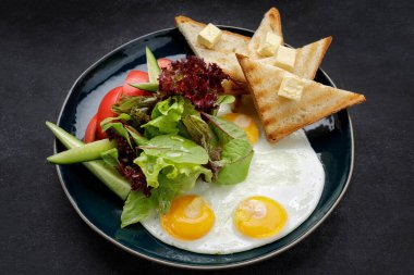 Kahvaltıda. Yağlı ve sebzeli kızarmış yumurta, salatalık ve domates. Karanlık bir tabakta, karanlık bir arka planda.