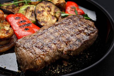 Sulu biftek ve ızgara sebze, tabakta baharatlar, koyu bir arka planda.