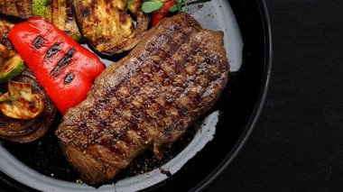 Sulu biftek ve ızgara sebze, tabakta baharatlar, koyu bir arka planda.