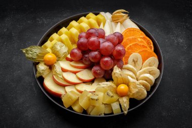 Tabakta dilimlenmiş meyve, tatlı. Karanlık bir arka planda. Mesajlaşmak için yer var. Üzüm, muz, portakal, armut, elma ananası