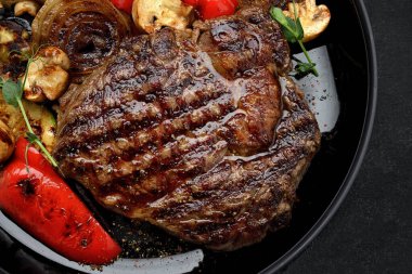 Fırında sebze ve mantarlı ızgara biftek, siyah bir tabakta, arka planda.