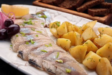 Tuzlu ringa balığı, patates, soğan, zeytin, limon ve tost, koyu bir arka planda.