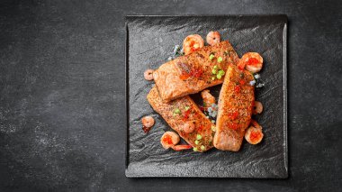 Kızarmış somon bifteği, karides ve kırmızı havyar, siyah bir tabakta, arka planda.