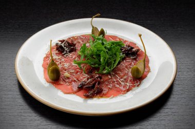 Dana fileto carpaccio, roka ve kapari, bir tabakta, koyu bir arka planda.